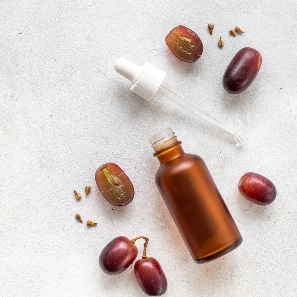 A small brown bottle next to a dropper top surrounded by re grapes on a white background.